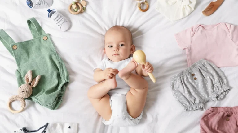 Newborn baby lying on a white bed holding a wooden rattle, surrounded by baby essentials including clothes, shoes, toys, and grooming items.