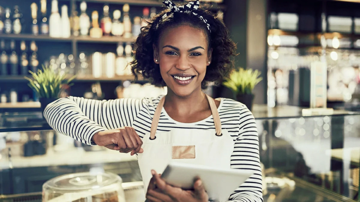 Smiling young small business owner wearing an apron standing in front of the counter of a trendy cafe using a digital tablet.