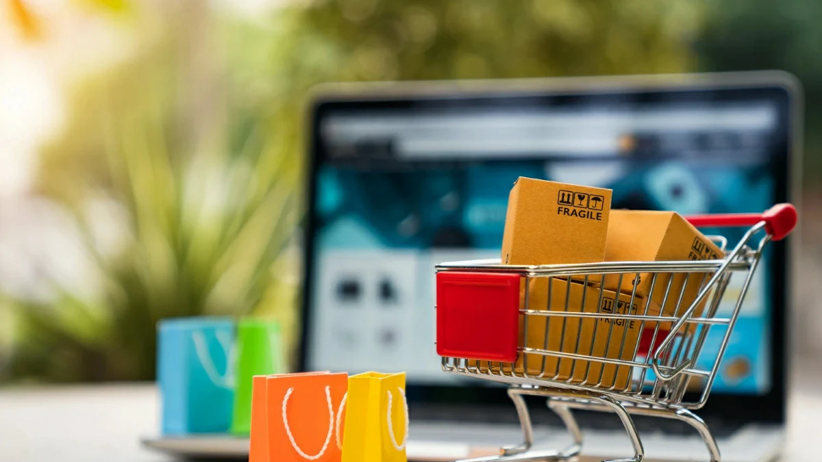 Product packages in a shopping cart next to a laptop displaying an online store, representing e-commerce, online business, and delivery services.