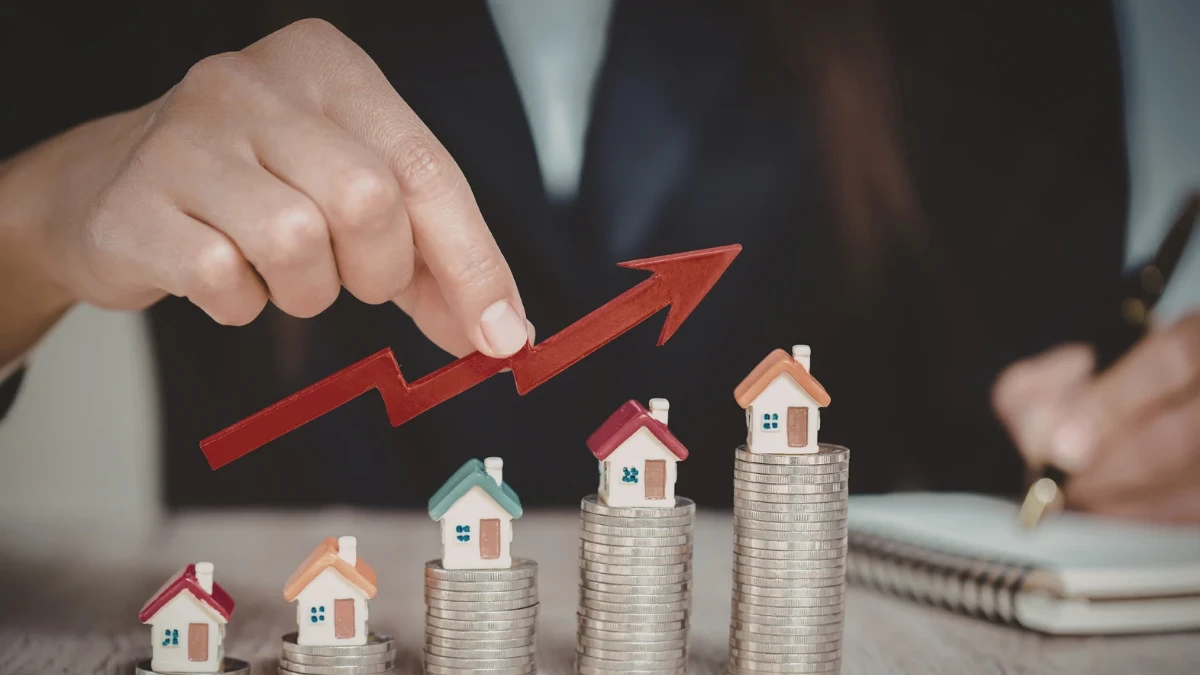Hand pointing to a red upward arrow over stacks of coins with small house models, symbolizing investing in real estate growth.
